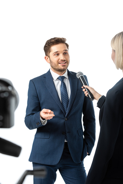 smiling businessman pointing with finger during interview with anchorwoman isolated on white, blurred foreground - Photo, Image
