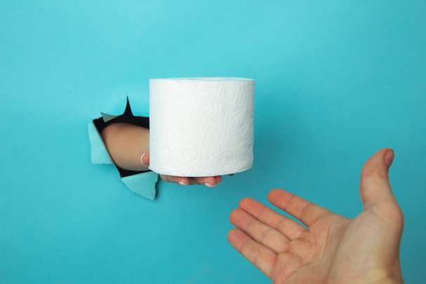 Mano femenina fuera del agujero en el papelero, sosteniendo un rollo de papel higiénico. Aislar sobre fondo azul - Foto, imagen