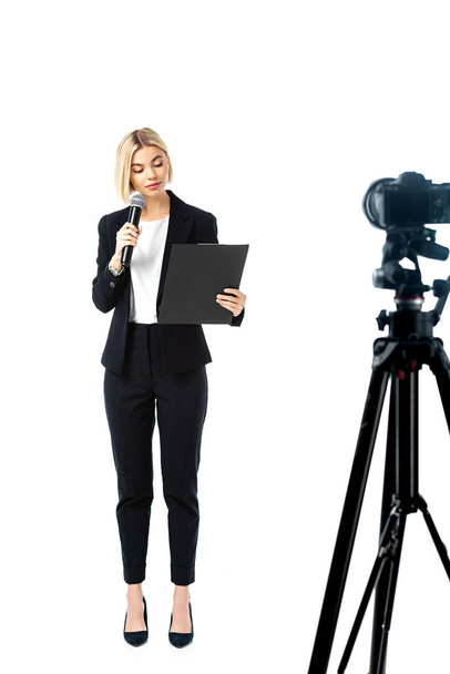 full length view of blonde news anchor in black suit near digital camera on white, blurred foreground - Photo, Image