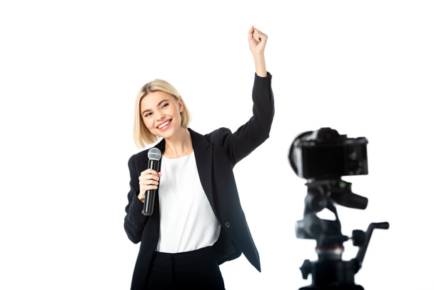 веселый вещатель с микрофоном, показывающий победный жест рядом с цифровой камерой на размытом переднем плане, изолированном на белом - Фото, изображение