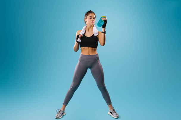 A young girl with an athletic physique drinks a protein cocktail while resting between exercises. Blue background and lots of empty space for advertising. - Valokuva, kuva