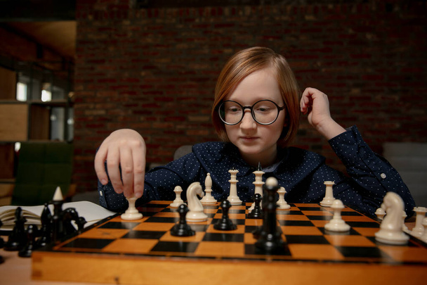 Gözlüklü kızıl saçlı çocuk evde oturmuş tek başına satranç oynuyor.. - Fotoğraf, Görsel