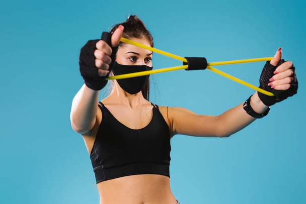 A young girl in sportswear and a black protective mask stretches a tourniquet for arm exercises with her hands. Blue background. - Φωτογραφία, εικόνα