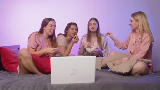 Τέσσερις χαρούμενες όμορφες γυναίκες με πιτζάμες κάθονται στο κρεβάτι, τρώνε ποπ κορν και βλέπουν ταινία στο μπάτσελορ πάρτι. - Πλάνα, βίντεο