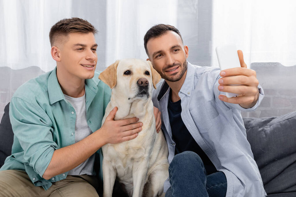 cheerful same sex couple smiling and taking selfie with labrador - Photo, Image