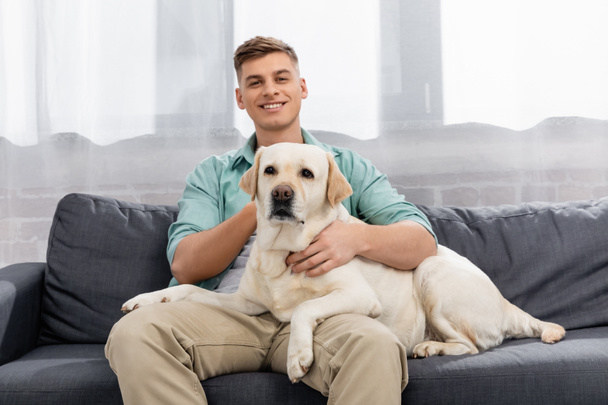 cheerful man sitting on couch and cuddling labrador - Foto, afbeelding