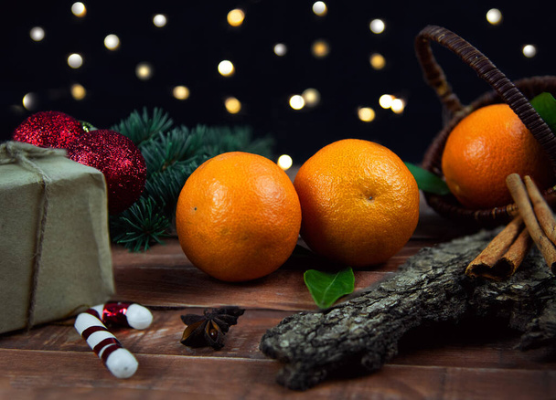 Μανταρίνια βρίσκονται σε μια καφέ ξύλινη σανίδα σε μαύρο φόντο, με τυλιγμένα δώρα, κλαδιά χριστουγεννιάτικου δέντρου και κόκκινα παιχνίδια χριστουγεννιάτικο δέντρο με ένα ραβδί κανέλας και γλυκάνισο αστέρι - Φωτογραφία, εικόνα