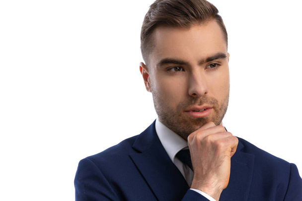 portrait of thoughtful young businessman in navy blue suit looking to side, holding hand to chin and thinking, standing isolated on white background in studio - 写真・画像
