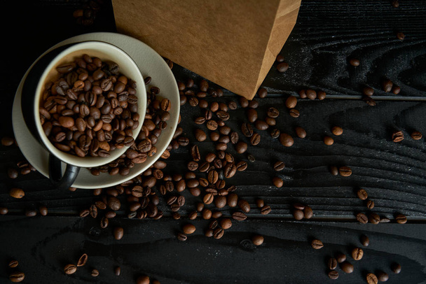 Κεραμικό κύπελλο γεμάτο με καβουρδισμένους μαύρους κόκκους καφέ, φωτογραφημένο σε μαύρες ξύλινες σανίδες με χάρτινη σακούλα και διάσπαρτους κόκκους καφέ στο σκοτεινό τραπέζι. - Φωτογραφία, εικόνα