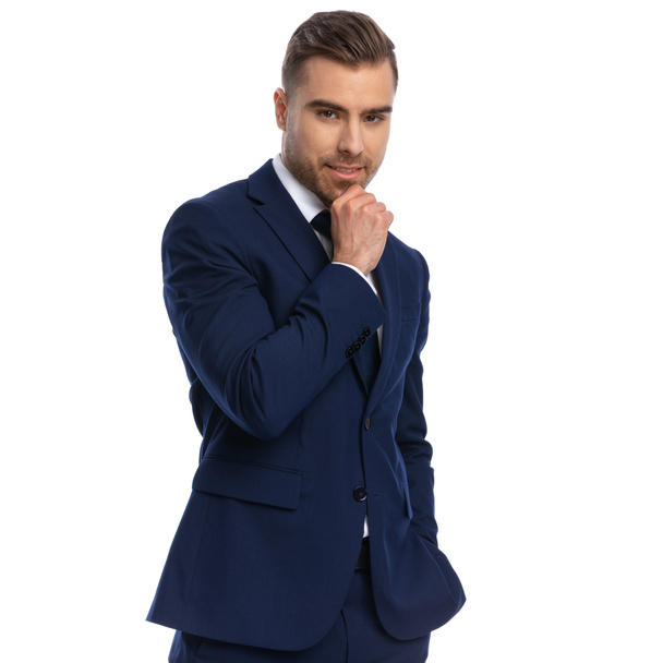 Porträt eines sinnlich eleganten Mannes im marineblauen Anzug, der die Hand ans Kinn hält und lächelnd auf weißem Hintergrund im Studio steht - Foto, Bild