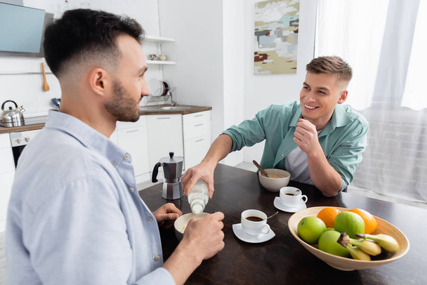 happy homosexual man pouring milk in bowl with corn flakes near husband  - Foto, Imagen