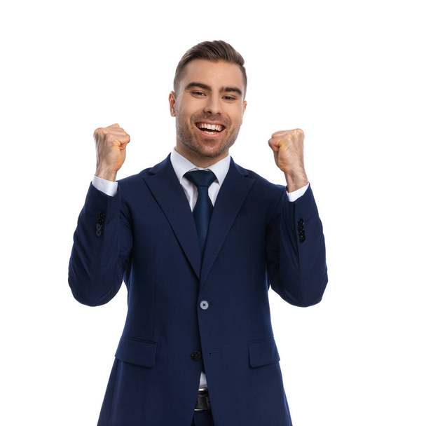 proud elegant man in navy blue suit holding fists in the air and celebrating victory, laughing and having fun, standing isolated on white background in studio, portrait - Фото, изображение