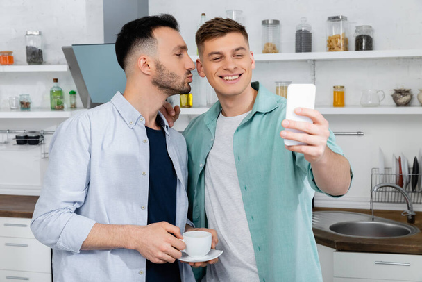 happy man pouting lips while kissing cheek of smiling husband taking selfie at home - Photo, Image