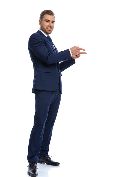 aantrekkelijke man met baard in marine blauw pak glimlachend, wijzend vingers naar opzij, poseren en staan geïsoleerd op witte achtergrond in studio, full body - Foto, afbeelding