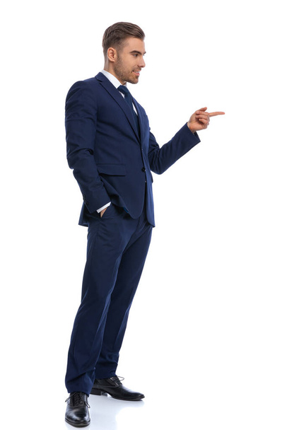 side view of elegant fashion model in navy blue suit pointing finger to side and flirting, holding hand in pocket and standing isolated on white background in studio, full body - Valokuva, kuva