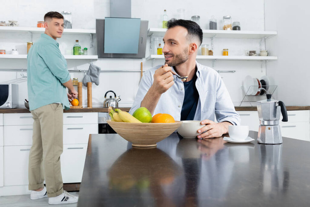 Χαρούμενος ομοφυλόφιλος άντρας που τρώει κορν φλέικς κοντά στο σύζυγο σε θολή φόντο - Φωτογραφία, εικόνα