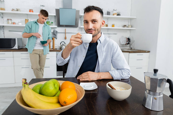 šťastný homosexuál pití kávy poblíž manžela na rozmazaném pozadí - Fotografie, Obrázek