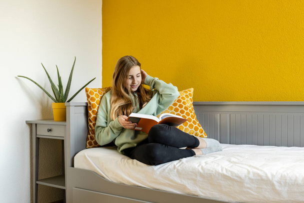 Young blonde teenage girl reading a book at home in front of a yellow wall - Zdjęcie, obraz