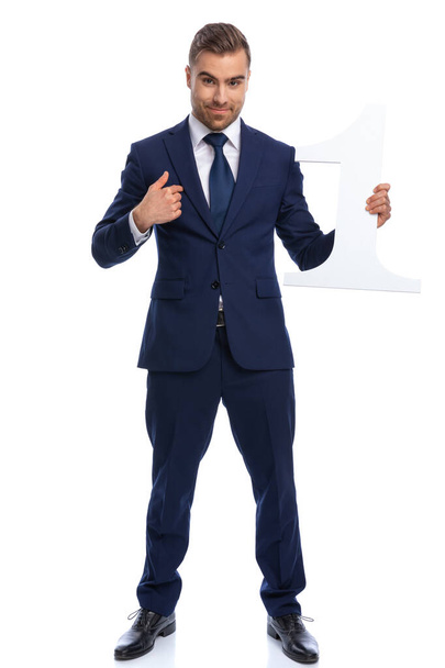 proud young businessman in navy blue suit pointing figer to himself, holding number one sign and smiling, standing isolated on white background in studio - Photo, Image