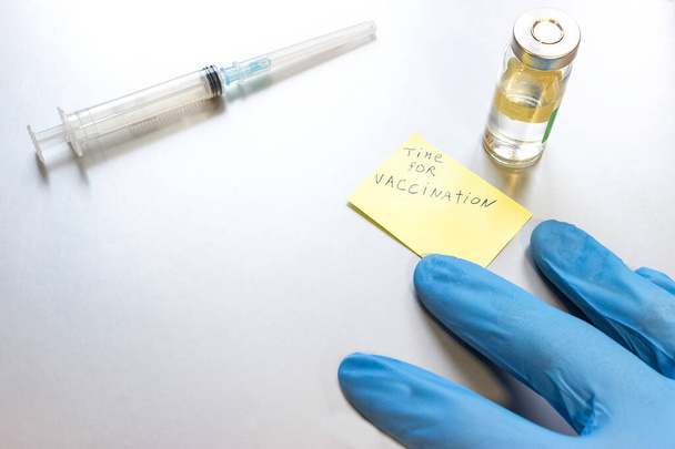 En esta foto Los médicos de la mano, la jeringa y la vacuna contra el coronavirus están sobre la mesa. concepto de vacunación gratuita en el mundo en 2021. la inscripción en la nota - tiempo para la vacunación. - Foto, Imagen