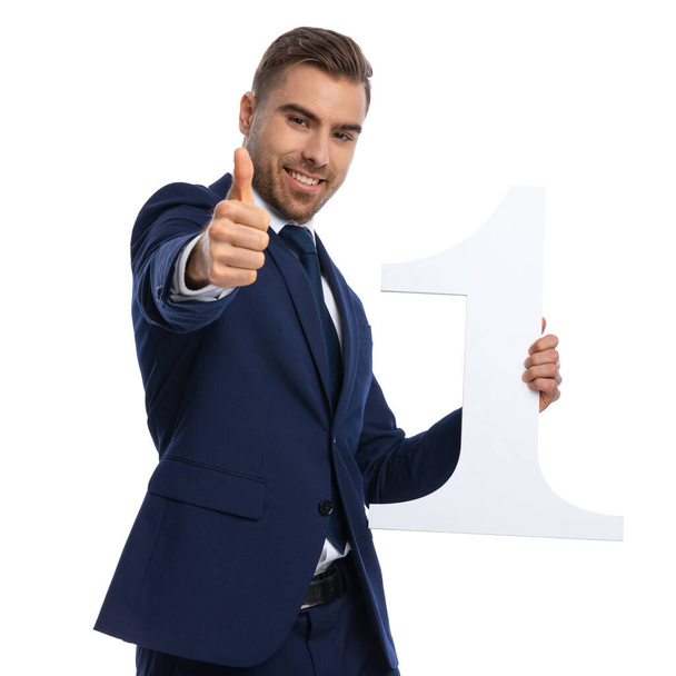 happy young businessman in elegant navy blue suit holding number one sign and making thumbs up gesture, standing on white background in studio - Photo, Image