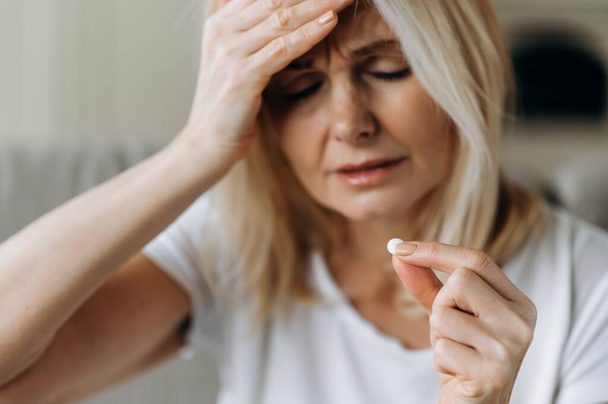 Upset unhealthy caucasian mature woman out of focus sitting at home on sofa drinking pain relieving pill for headache with eyes closed - Foto, Imagem