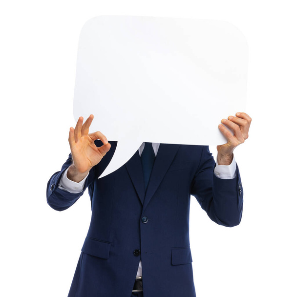 συνεσταλμένος νεαρός επιχειρηματίας σε μπλε σκούρο εξώφυλλο με φούσκα ομιλίας και κάνοντας το ok sign να στέκεται απομονωμένο σε λευκό φόντο στο studio - Φωτογραφία, εικόνα