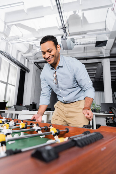 Cheerful indian businessman playing table soccer on blurred foreground in office  - Foto, Bild