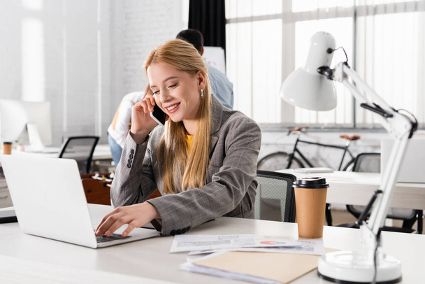 Smiling businesswoman using laptop and talking on smartphone near documents and lamp in office  - Foto, Imagem