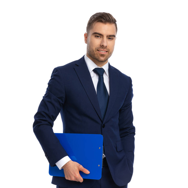 attractive unshaved guy holding hand in pocket, holding clipboard and smiling, posing on white background in studio - Fotoğraf, Görsel