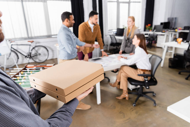 Uomo di consegna che tiene scatole di pizza vicino a uomini d'affari su sfondo sfocato in ufficio  - Foto, immagini