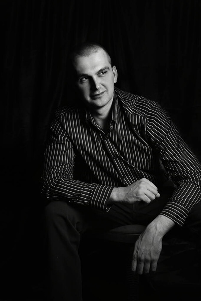 Zwart-wit portret van een man in de studio, zittend met zijn handen zijn gebald in vuisten, kijken aan de zijkant - Foto, afbeelding