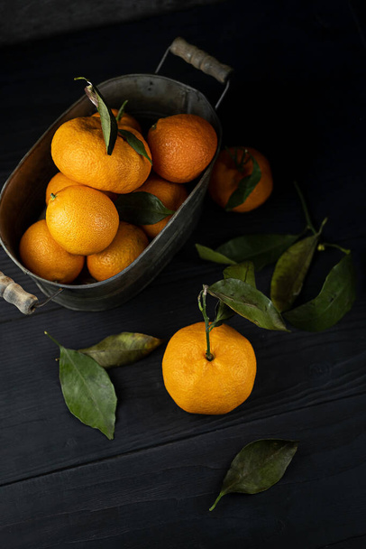 Several tangerines with leaves in a container lie on a black table against a dark background. Rustic style copyspace. - Φωτογραφία, εικόνα