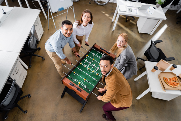 Vista aérea de la alegre gente de negocios multiétnicos jugando futbolín cerca de la pizza en la oficina  - Foto, imagen