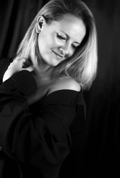 Retrato en blanco y negro de una mujer rubia tierna y sensual en el estudio, de pie con el cuerpo medio doblado en una camisa negra con hombros desnudos. Su mirada se dirige hacia el lado hacia abajo, una mano cerca del pecho sostiene la camisa - Foto, Imagen