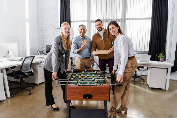 Sonriendo gente de negocios interracial con pizza mirando a la cámara cerca de futbolín en la oficina  - Foto, imagen