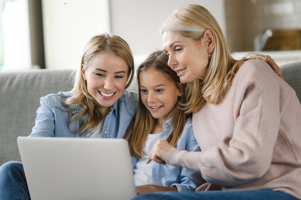 Happy caucasian grandmother, daughter and granddaughter spending time together, sitting at home on the sofa using laptop, browsing network, shopping online, smiling - Fotoğraf, Görsel
