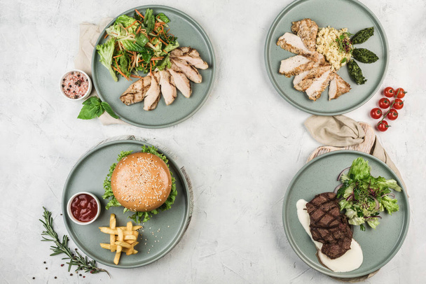 Several warm dishes with chicken, turkey, beef fillets and pork cutlet burgers in different plates served by the chef on a light background, top view with a copispace. flat lay. Restaurant food. - Photo, Image