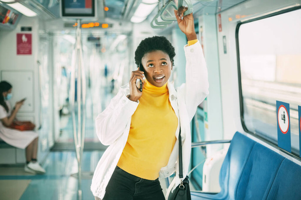 Hymyilevä nuori afrikkalainen nainen pitää kiinni kaiteesta ja puhuu puhelimessa, seisoo metrojunassa päivittäisen työmatkasi aikana.. - Valokuva, kuva