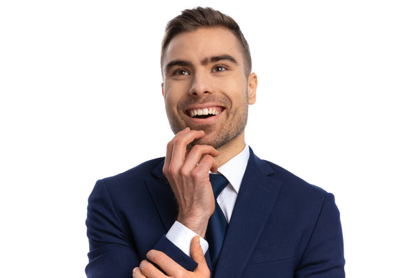 portrait of fascinated bearded man in elegant navy blue suit holding hand to chin, looking up and showing admiration, smiling and posing on white background in studio - Fotoğraf, Görsel
