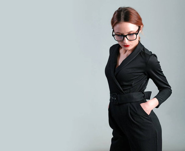 Elegant business woman in black classic jumpsuit and glasess posing over gray background. - Photo, Image