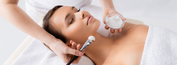 esteticista segurando escova cosmética e recipiente com máscara facial perto do cliente na mesa de massagem, banner - Foto, Imagem