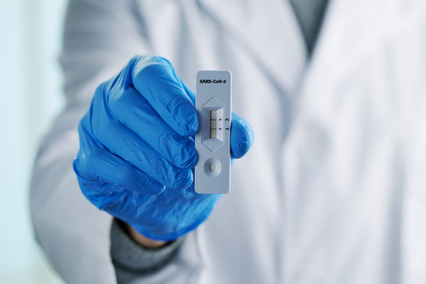 closeup of a healthcare worker, wearing a white coat and blue surgical gloves, holding a covid-19 test device showing a positive result in his hand - 写真・画像