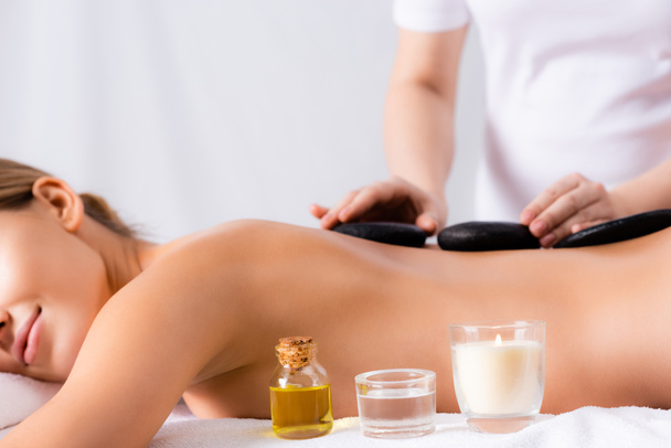 aromatic candle and bottle with oil near young woman getting hot stone massage on blurred background - Photo, Image
