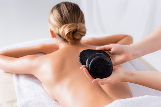 vista ritagliata del massaggiatore in possesso di pietre calde nere vicino al cliente nel salone spa  - Foto, immagini