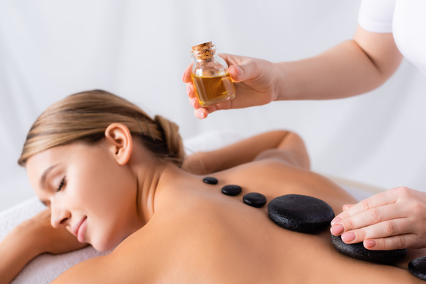 masseur holding fles met olie in de buurt klant het krijgen van hete steen massage - Foto, afbeelding