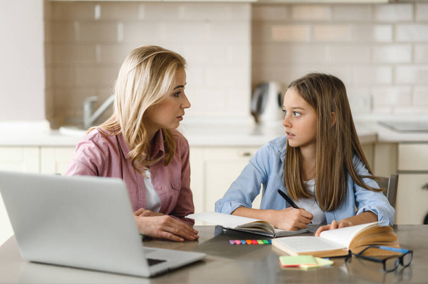 Distance learning. Mom helps her daughter do her homework, they sit at the table at home, using a laptop, books and a notebook. Homeschooling - Photo, Image