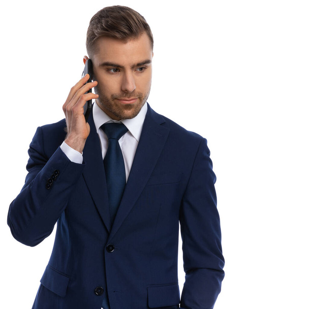 portret seksownego, nieogolonego biznesmena w granatowym garniturze spoglądającego w bok i rozmawiającego przez telefon, stojącego na białym tle w studio - Zdjęcie, obraz