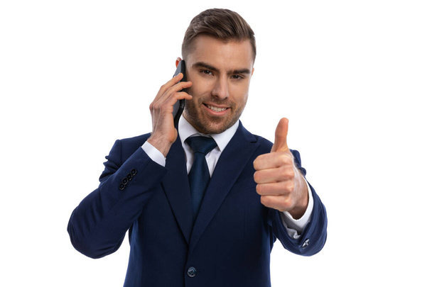 heureux jeune homme en costume bleu marine souriant, parlant au téléphone et faisant le pouce vers le haut geste, debout isolé sur fond blanc en studio - Photo, image