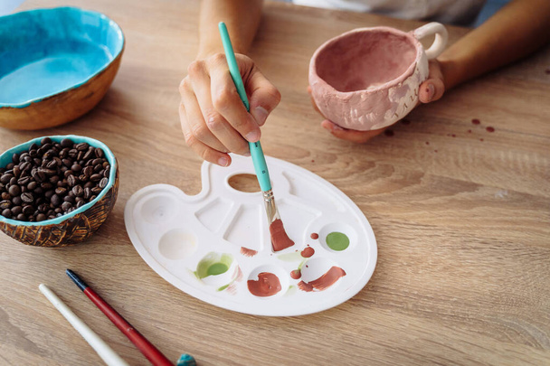 木製のテーブルの上のパレットに絵筆を浸し手。粘土陶器生産の仕上げ製造プロセス。独占手作り製品ターコイズとロイヤルブルーのボウル - 写真・画像